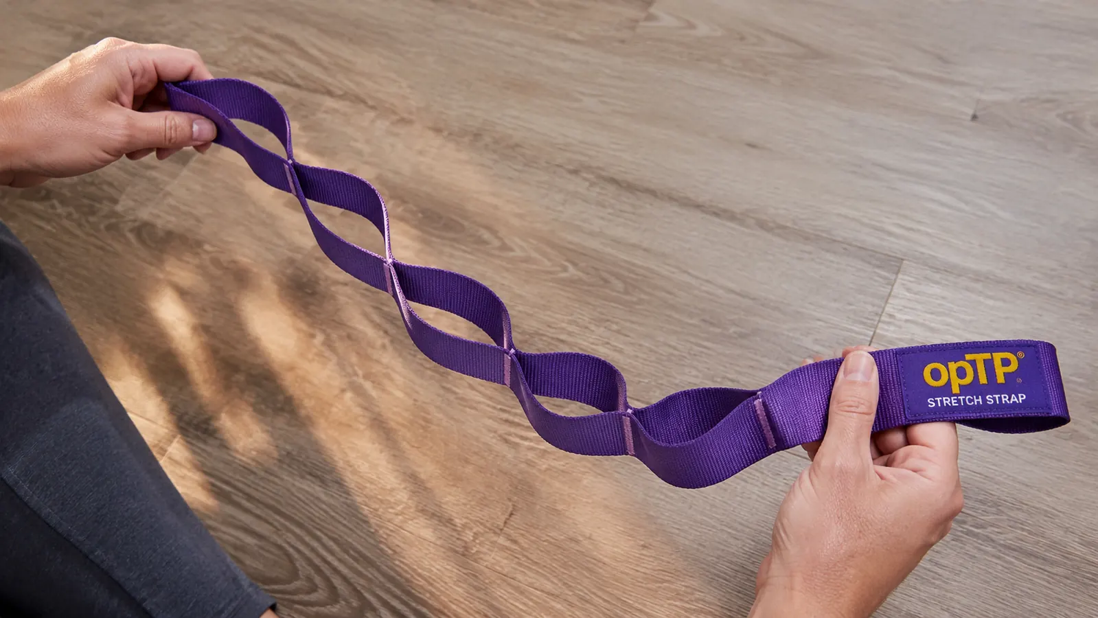 Close-up of hands holding the OPTP Stretch Out Strap, showing the multiple webbed loops along its length on a wood surface
