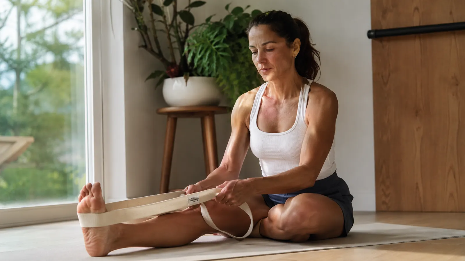 Person sitting on a yoga mat holding the OPTP Stretch Out Strap around the arch of their foot, hamstring stretch in progress, bright morning light from a window